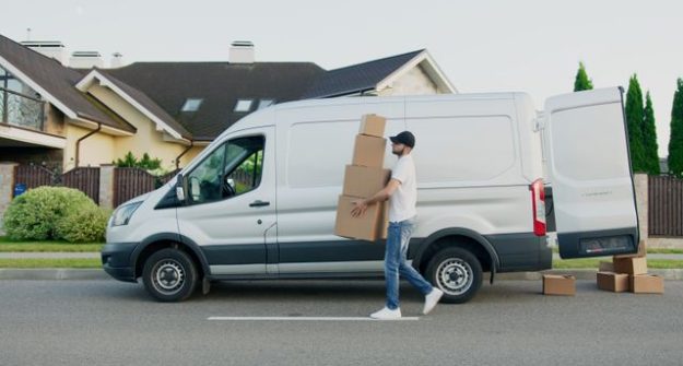Man unloading cardboard boxes from a delivery van in a suburban neighborhood street.